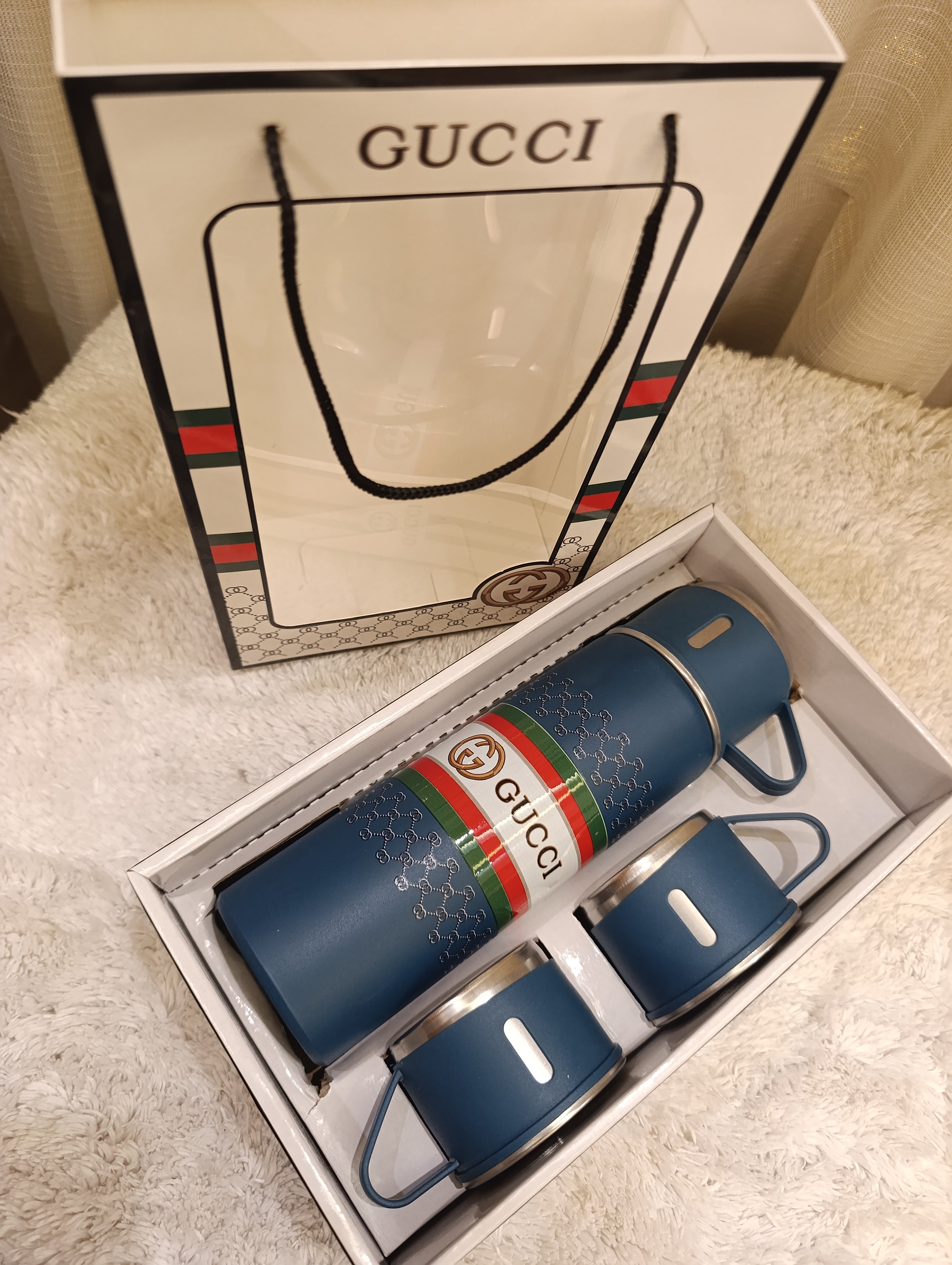 A Gucci-branded blue insulated bottle and two matching cups presented in an open box, with a Gucci shopping bag standing behind on a soft white faux-fur surface.