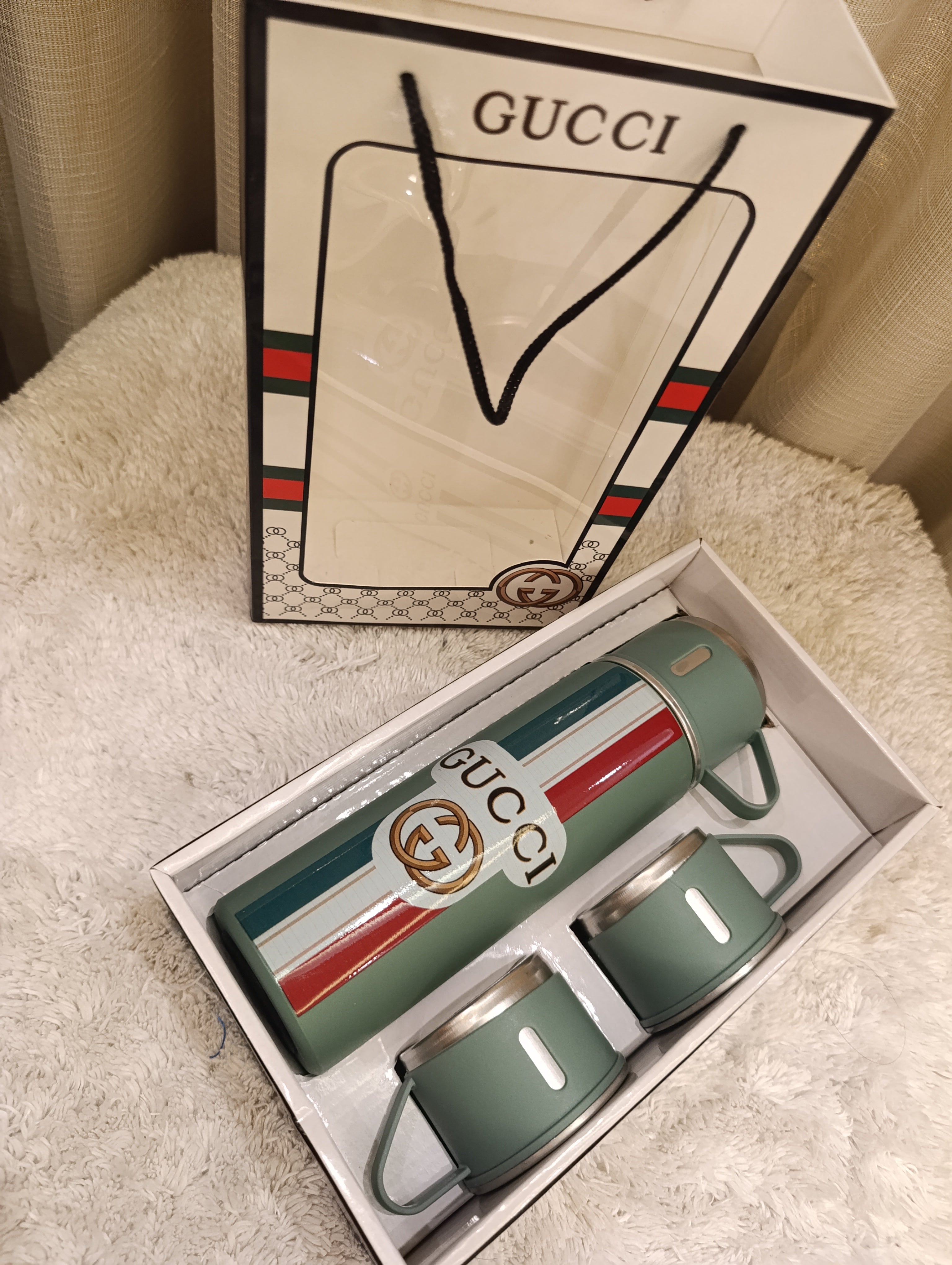 Gucci travel set featuring a green insulated bottle and two matching cups in a white box, with a branded Gucci shopping bag standing behind on a cream textured surface.