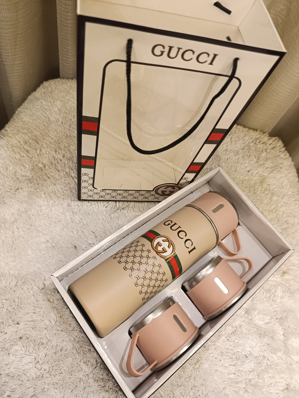 A Gucci-branded insulated travel set displayed in an open box, featuring a beige water bottle with a green and red stripe pattern and two matching beige cups with stainless steel lids, alongside a branded Gucci shopping bag on a cream textured surface.